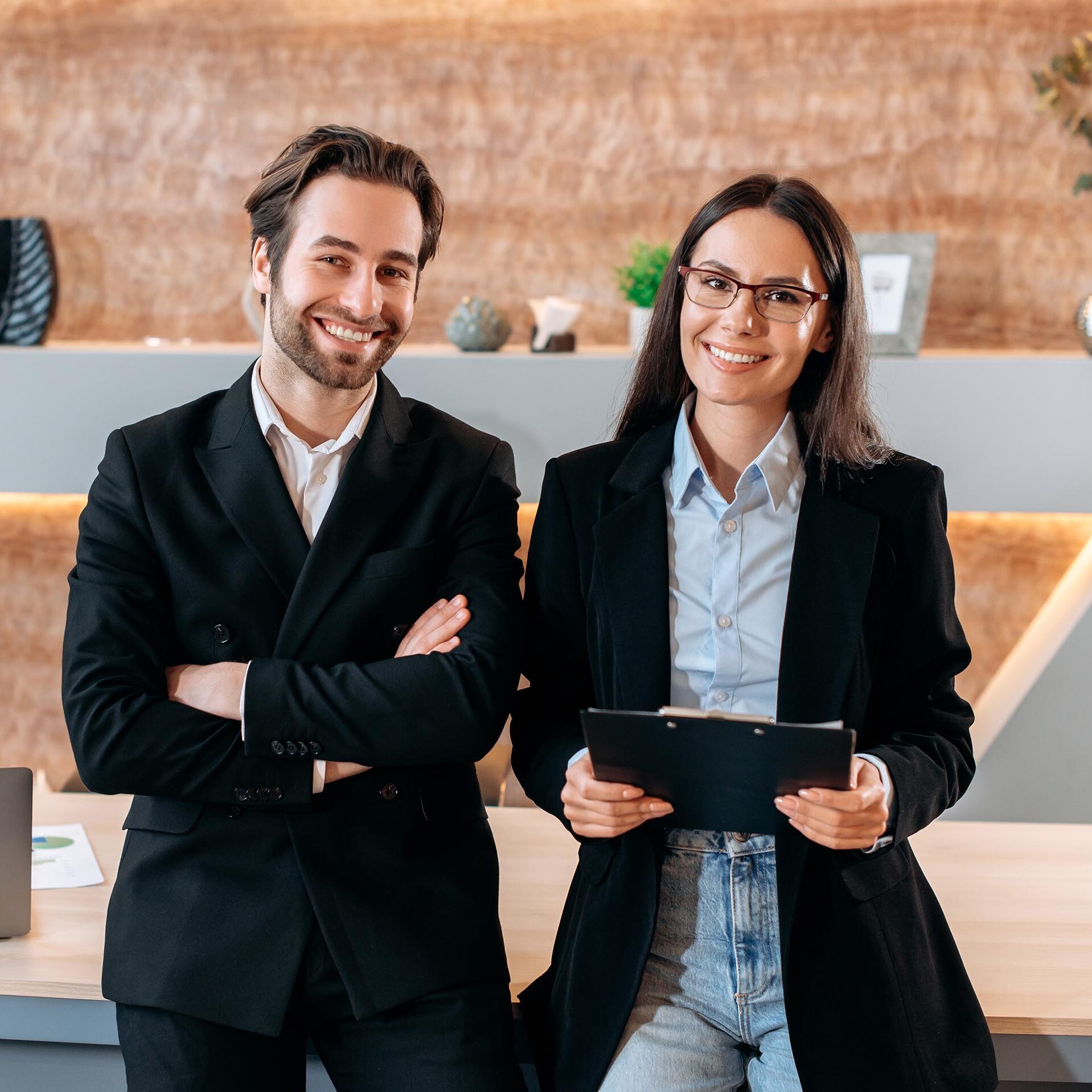 Portrait of two caucasian confident successful company employees, business colleagues, a man and a woman, stand near the desktop in a modern office, look at the camera, smile friendly Portrait of two caucasian confident successful company employees, business colleagues, a man and a woman, stand near the desktop in a modern office, look at the camera, smile friendly